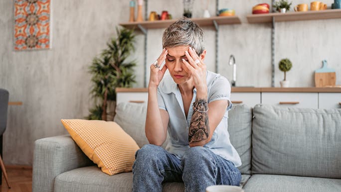 A middle-aged woman with short grey hair and a tattoo on her arm is sitting on the sofa, holding her head with both hands, possibly experiencing a headache due to dehydration