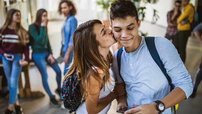 A teenage girl kisses her crush on the cheek, there are other students in the background looking shocked. 