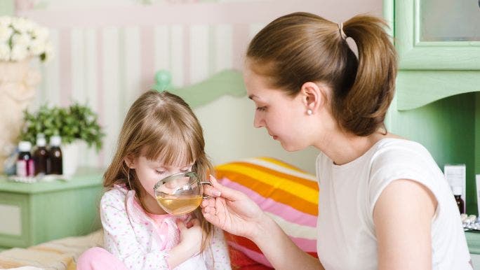 A young mother is offering a cup of hot tea to a sick young girl who is in bed