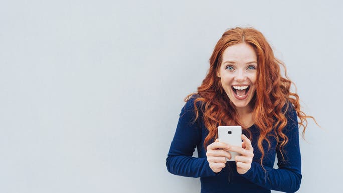 Vivacious young woman laughing at a good joke as she stands against a white wall with copy space holding a mobile