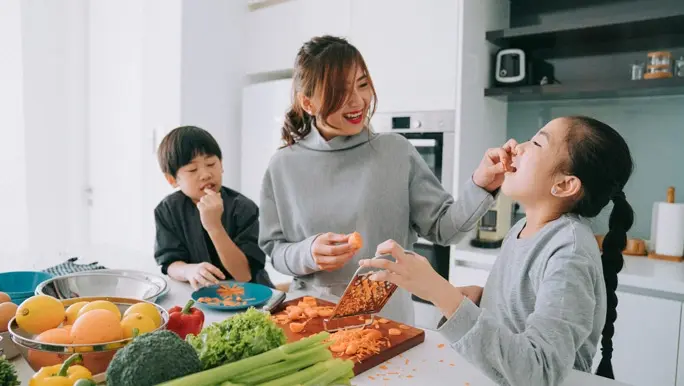 Mother enjoy preparing food at kitchen counter with her son and daughter feeding her daughter with shredded carrot stock