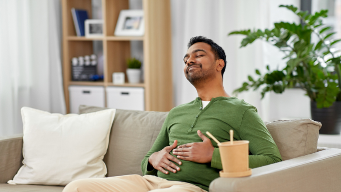 A young man with a dark complexion is sitting on a sofa smiling and holding his belly