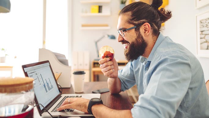 A young man with brown hair and a beard is eating an apple and smiling as he works on a laptop to find answers to mens health questions