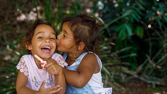 Two little girls sit at a metal picnic table, one is eating an ice cream and smiling as the other kisses her cheek. 
