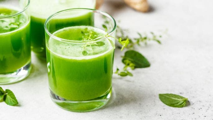 Two glasses of green juice are on a table with ginger and leaves in the background