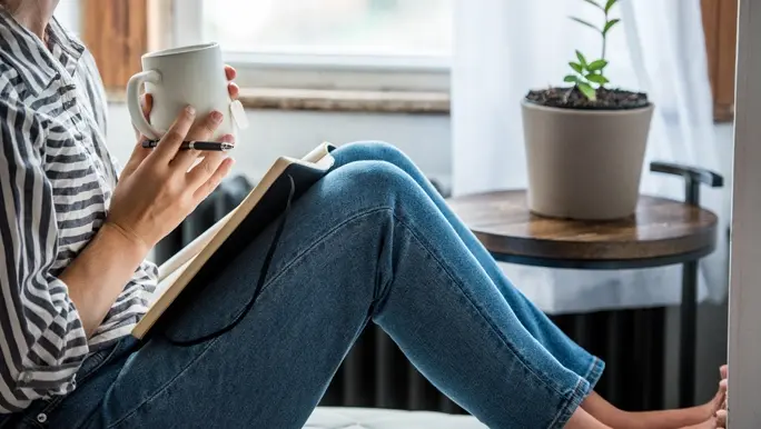 Women in jeans and striped shirt sitting by the window writing in a notebook and sipping tea from a white mug
