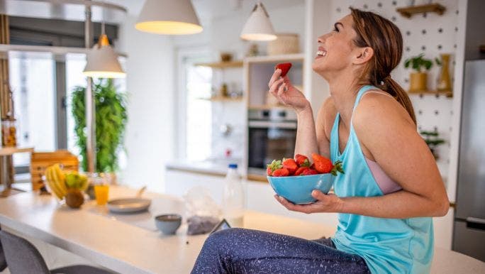 A young woman wearing activewear is sitting on a kitchen bench smiling as she eats a bowl of strawberries
