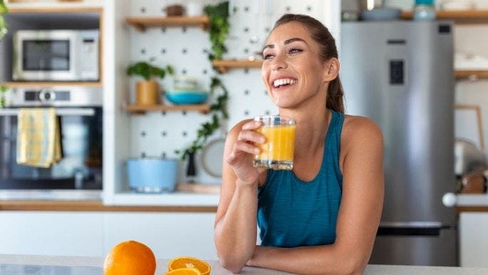 A woman sits at her kitchen bench drinking a glass of orange juice