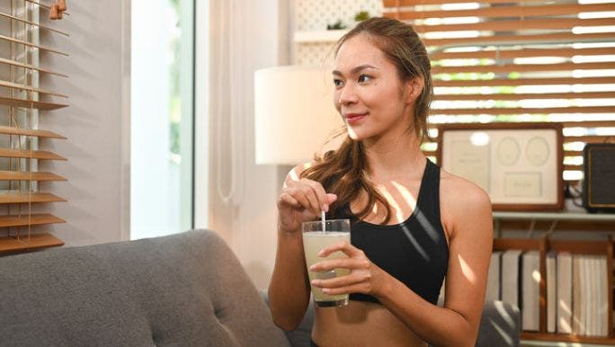 A young woman wearing activewear is smiling as she stirs a collagen supplement in a glass of water