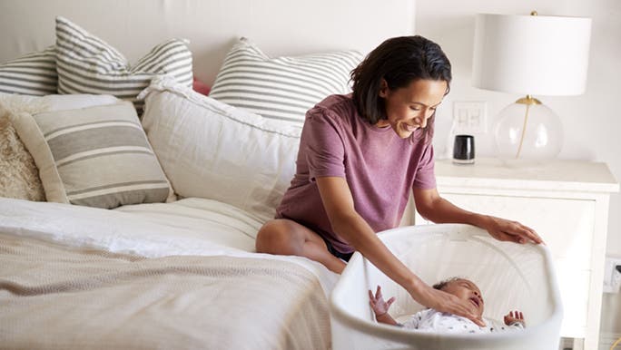 A lady sits cross legged on a plush bed, she is smiling and talking calmly to her baby who is laying in a bassinet, crying in their sleep.