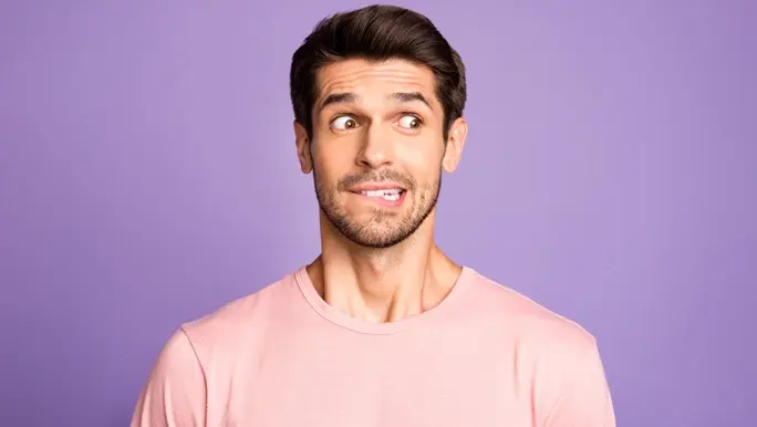 Man with a slightly embarrassed expression. He is wearing a pink t-shirt, standing in front of a purple background 