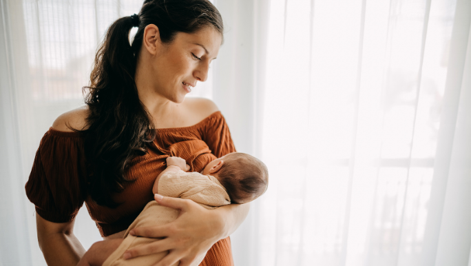A young brunette woman is standing in front of a curtained window and breastfeeding her baby