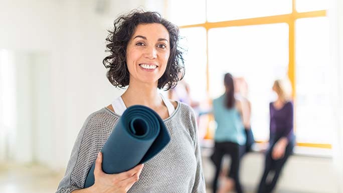 A woman walking into a pilates class smiles with a rolled up exercise mat and wonders what the pelvic floor is. 