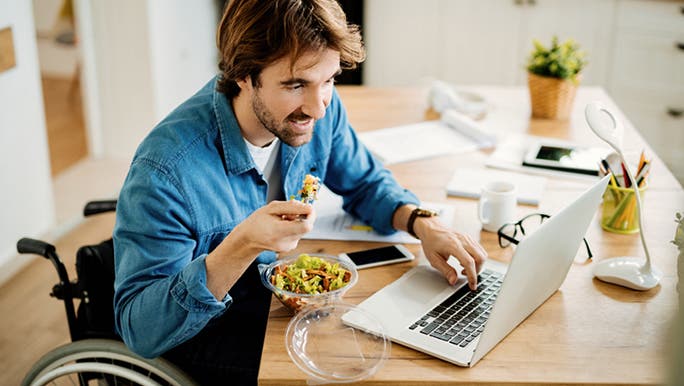 A young man in a wheelchair and wearing a blue shirt eating a salad while working on a laptop