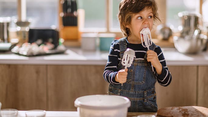Child standing at a kitchen bench licking icing off beaters. His parents are about to experience the effects of too much sugar. 