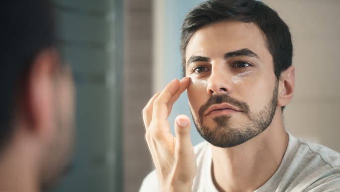 A man applies moisturiser underneath his eyes while looking into the mirror and wondering what the moisturiser does. 