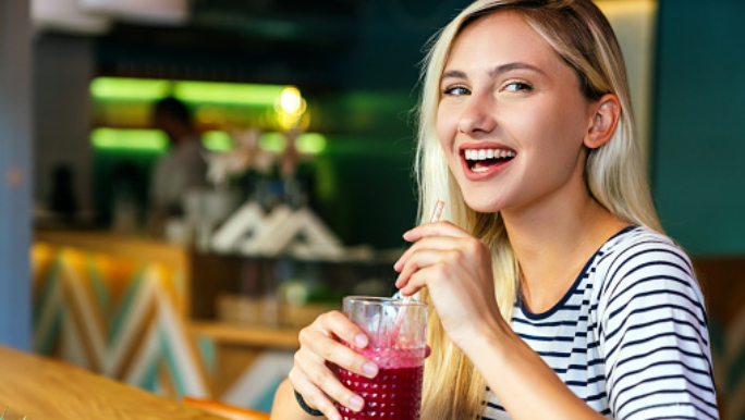 A young blonde woman is smiling and drinking beetroot juice