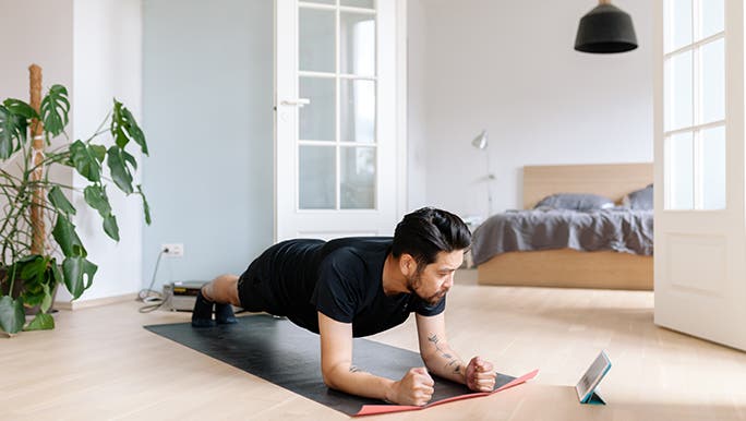 A man planks in his home in front of a monstera plant. It looks like he is following directions on an iPad. 