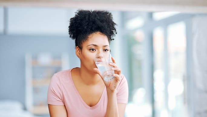 A woman drinks a glass of water because she hopes it will help to hydrate her skin. 