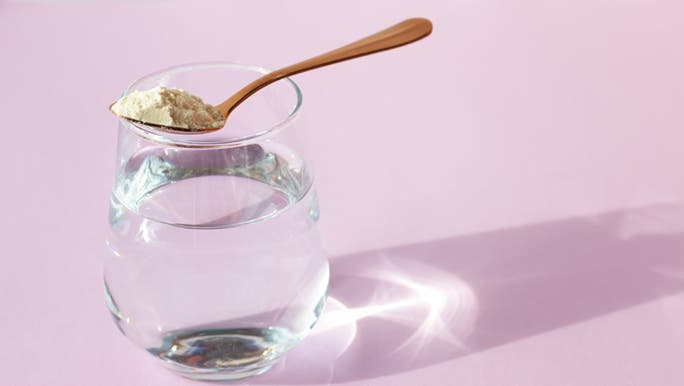 On a pink background, a bronze spoon filled with collagen powder perches on a glass of water.