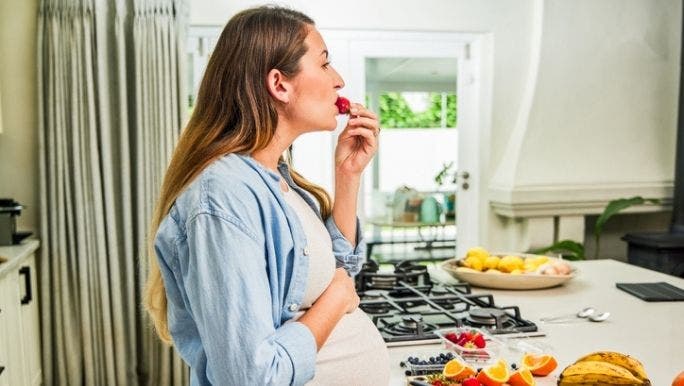A pregnant woman is standing in a kitchen eating a strawberry while holding her pregnant belly