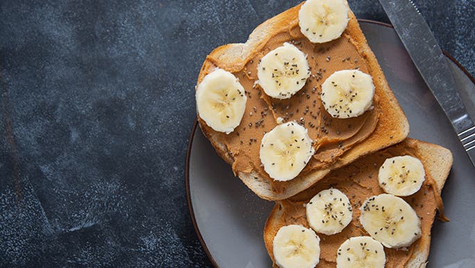 Two slices of peanut butter toast are on a plate, they have sliced banana and chia seeds on them. 
