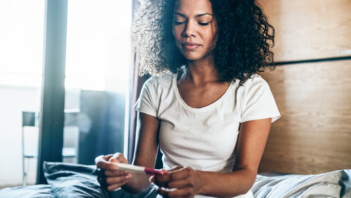A woman with dark curly hair and dark skin sits on a bed looking at an ovulation calculator for signs of ovulation