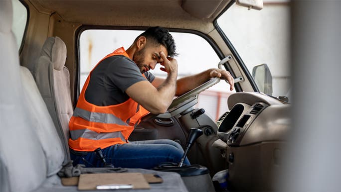A young man wearing a high-vis vest is sitting in a truck with his head in his hand looking tired