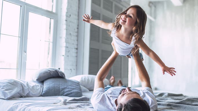 A man is lying on an unmade bed playfully holding a young girl up in the air 