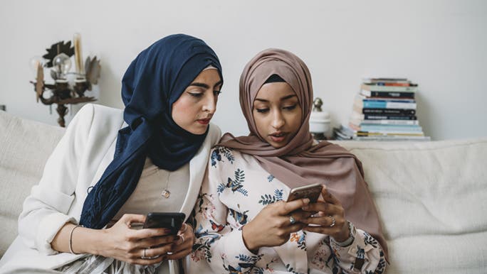 Two women sit on a white couch looking at social media on their phones. 