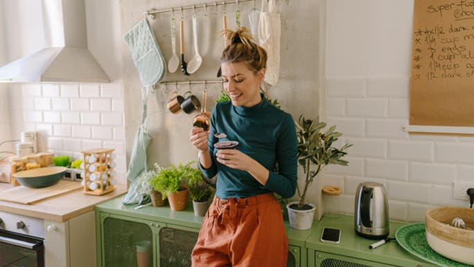 A young woman wearing a blue polo neck top and orange pants is enjoying mindful eating as she smiles and eats a snack in her kitchen