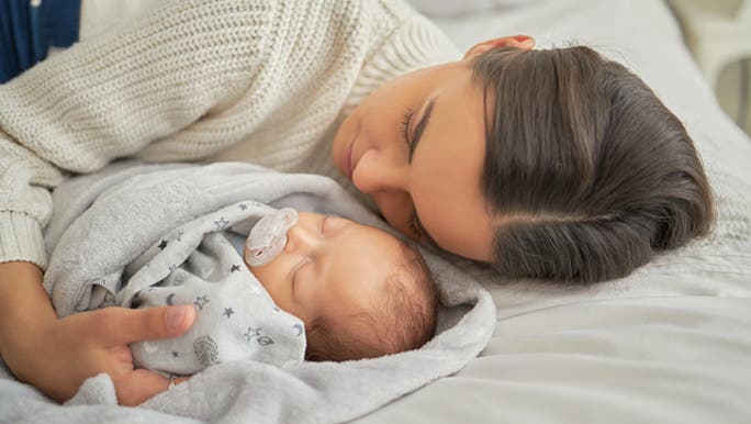 A woman in a white jumper has her arm around her newborn baby as they both lie on a bed resting