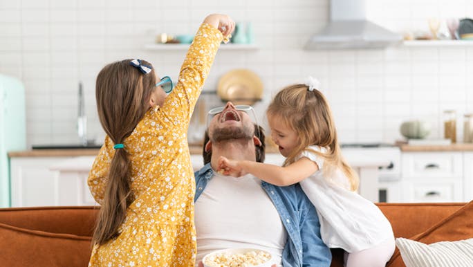 Two little girls on a couch are feeding a man popcorn, a great healthy snack for kids. 