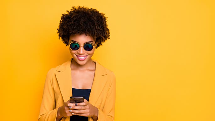 A lady in a yellow suit standing in front of a yellow wall smiles at her phone which she is viewing a social media platform on. 