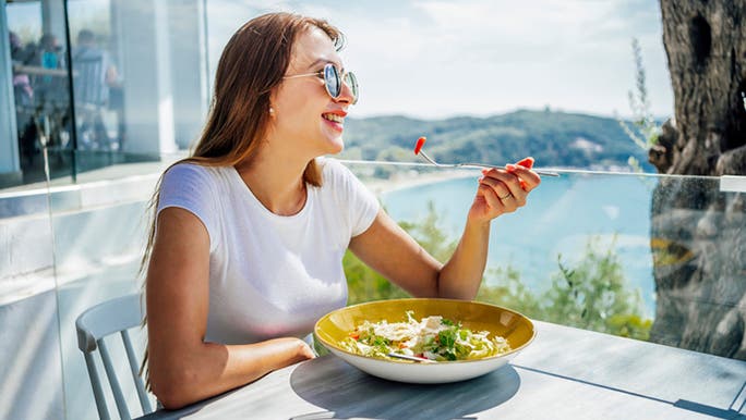 A young woman wearing sunglasses and a white T-shirt eating a Mediterranean salad on a balcony overlooking the water