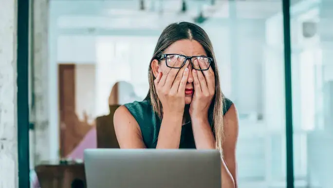 Woman has her head in her hands at work while she sits in front of her laptop experiencing work-related stress.