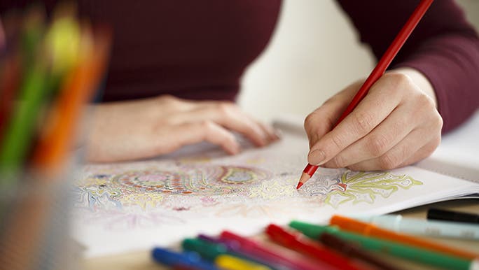 A close up image of a woman’s desk and arms, she is using a mindfulness colouring book. 
