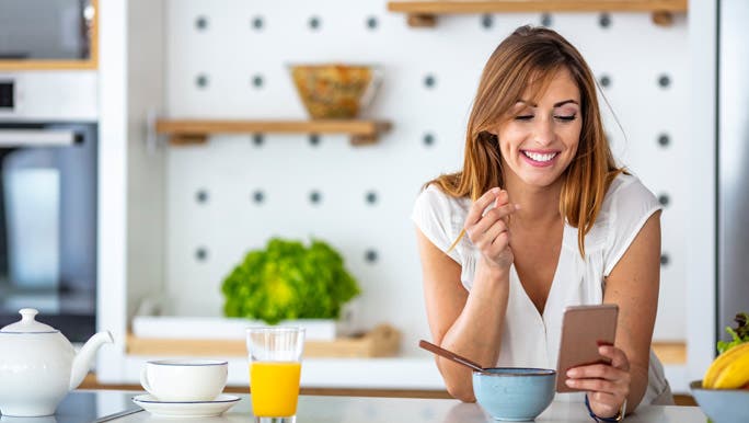 Woman snacking in her kitchen and looking at her smartphone