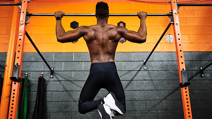 From behind, a muscly man is doing chin ups on a bar in front of a yellow and black wall, in the hopes of bulking up.