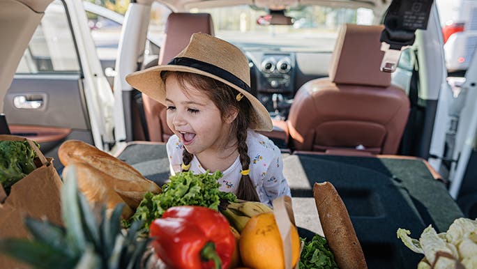 A toddler is in the boot of a car, excitedly looking at a box full of veggies. 