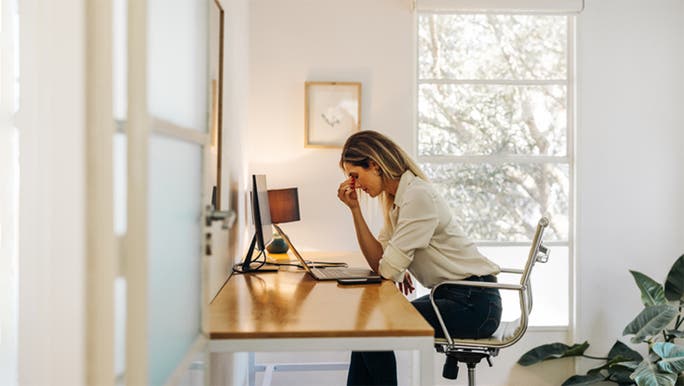 A young blonde woman is sitting at a computer desk touching her face and looking tired
