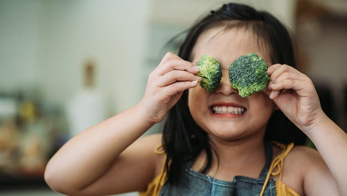 A little girl is holding broccoli in front of her eyes and smiling. She is choosing healthy food over junk food. 