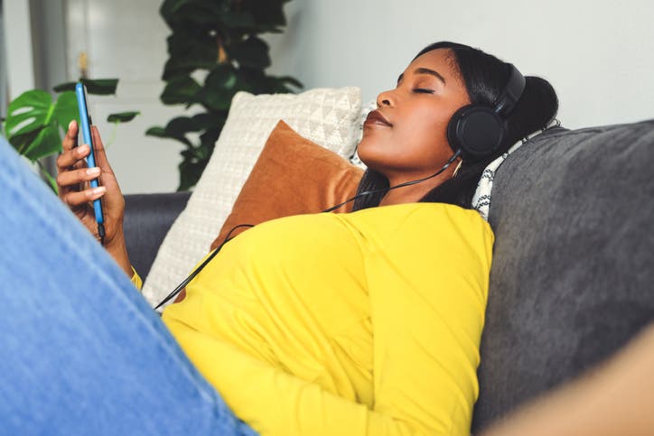 A dark-skinned woman with black hair is wearing a yellow top and sitting on a sofa while she listens to her smartphone on a set of headphones