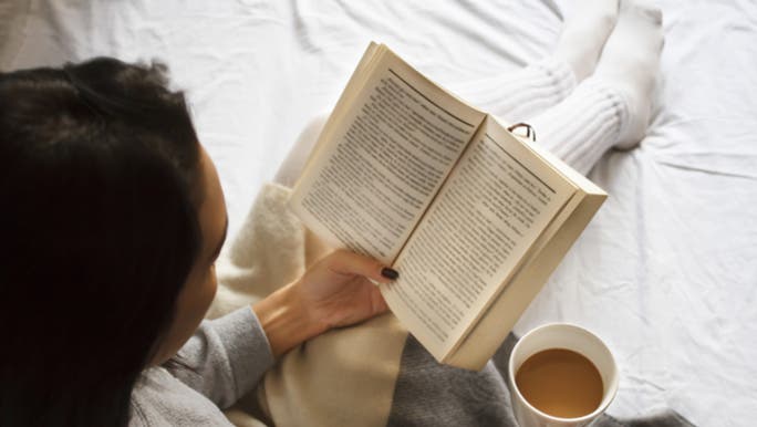 A woman is reading a book in bed with a cup of tea in her hand. She is avoiding screen time as it affects her sleep. 