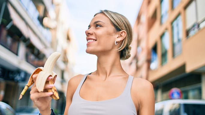 A young Caucasian woman is walking in a city wearing a singlet and eating a banana, which may be linked to gut health