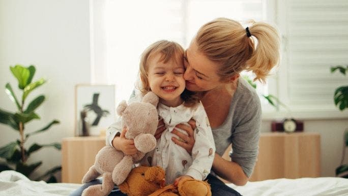 A young mother is hugging her young daughter and kissing her cheek while sitting on a bed