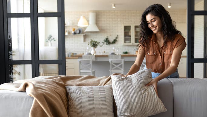 A young woman with long dark hair is smiling as she arranges cushions on a sofa
