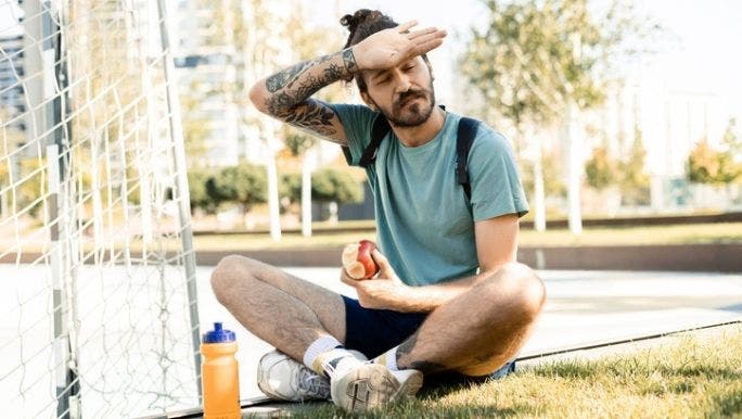 A young man with tattoos is sitting on the ground eating an apple and wiping sweat from his brow