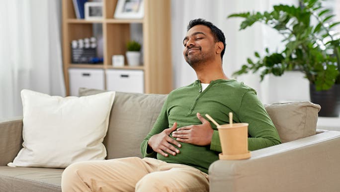 A man with dark hair and skin wearing a green jumper and sitting on a couch is smiling as he rubs his belly, thinking about the connection between gut health and mental health