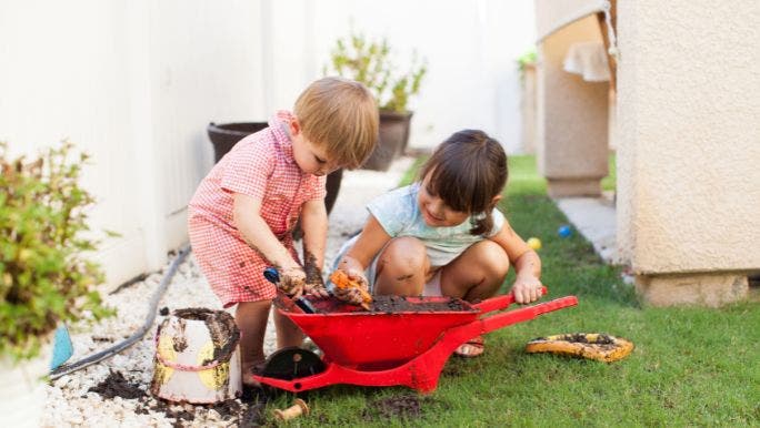 Two young children are playing with a red wheelbarrow in a garden